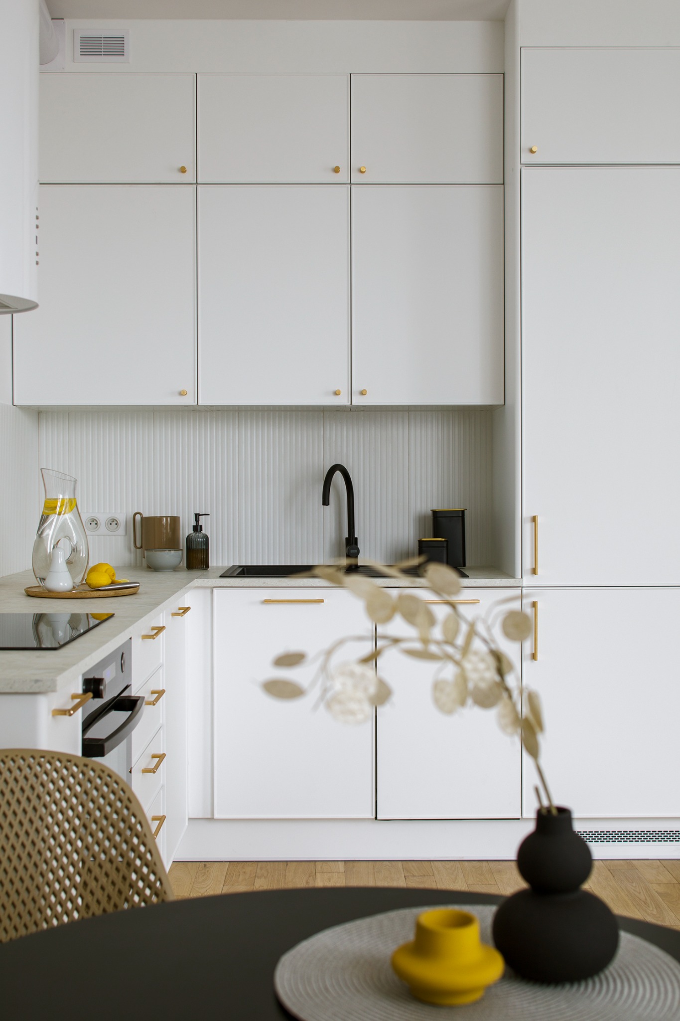 Minimalistyczna kuchnia w bieli z akcentami złota: armatura, uchwyty, detale dekoracyjne, czarny zlew i bateria, na blacie dzbanek z wodą i cytryną, wazon z suszem na stole.