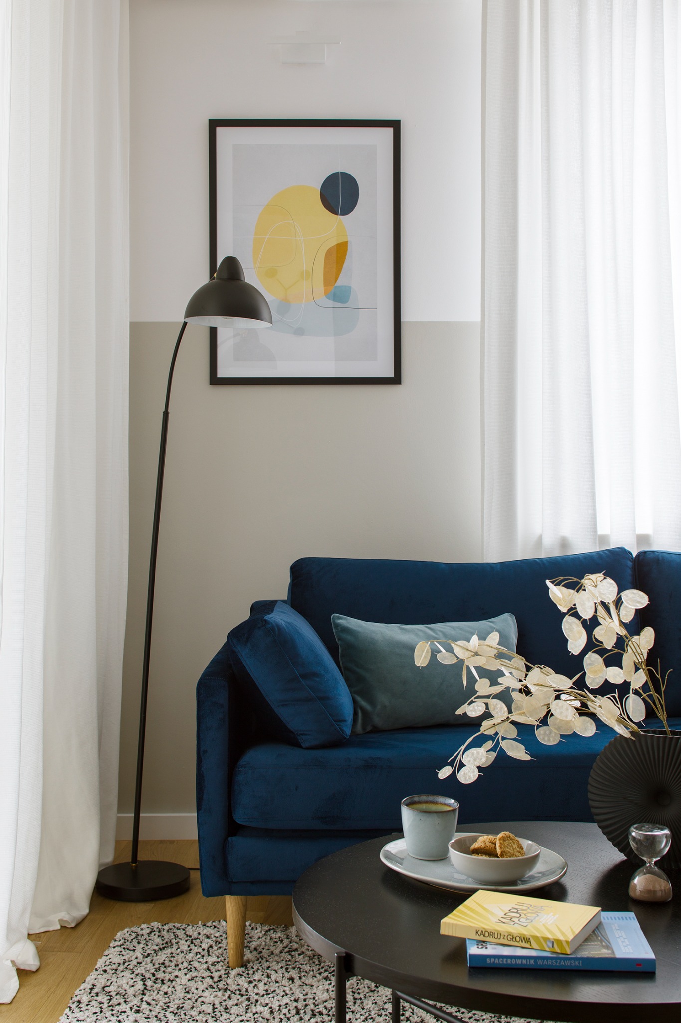 Niebieska welurowa sofa z poduszkami i dekoracją z suszonych roślin, czarny stolik kawowy z książkami i przekąskami, lampa podłogowa i abstrakcyjny obraz na ścianie w pokoju z białymi zasłonami.