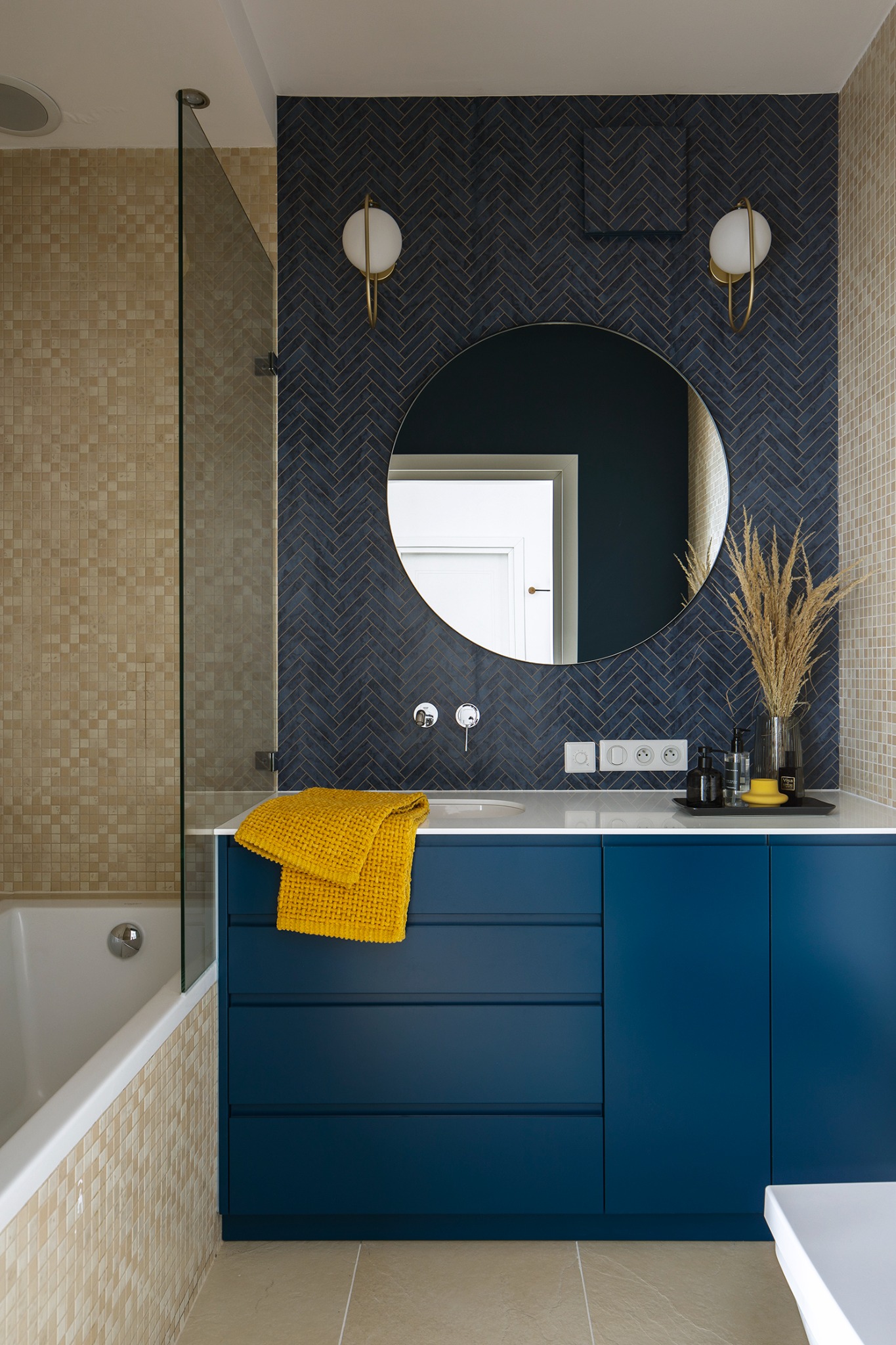Nowoczesna łazienka z niebieską zabudową szafek pod umywalką, okrągłym lustrem na ścianie z ciemnymi płytkami ułożonymi w jodełkę, beżowymi mozaikami na ścianach, żółtym ręcznikiem i szklanym...