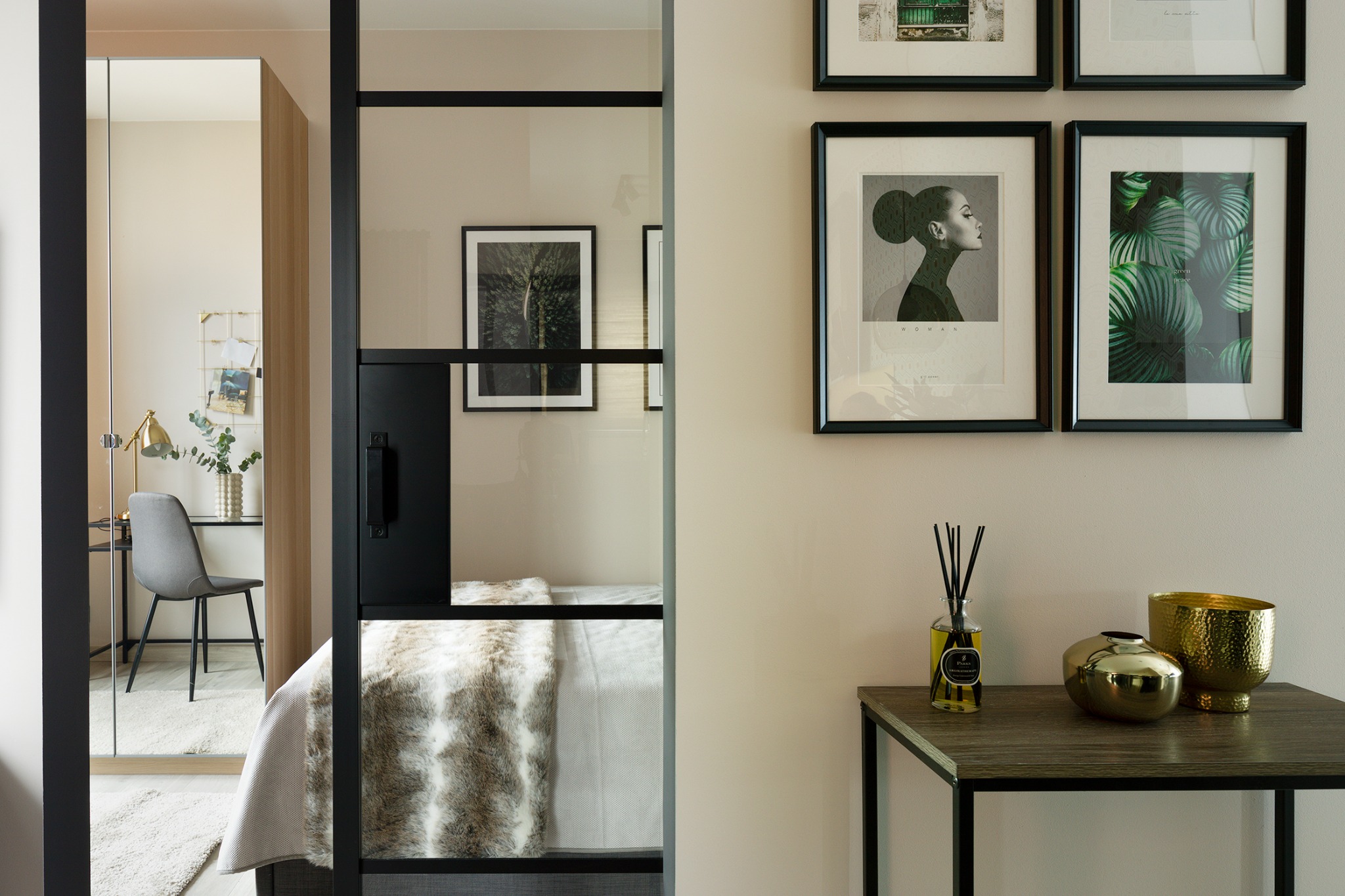 Sypialnia z wydzielonym miejscem do pracy, oddzielona szklaną ścianą z czarnymi szprosami; na ścianie galeria czarno-białych grafik, na stoliku dekoracyjny dyfuzor zapachowy i złote wazony.