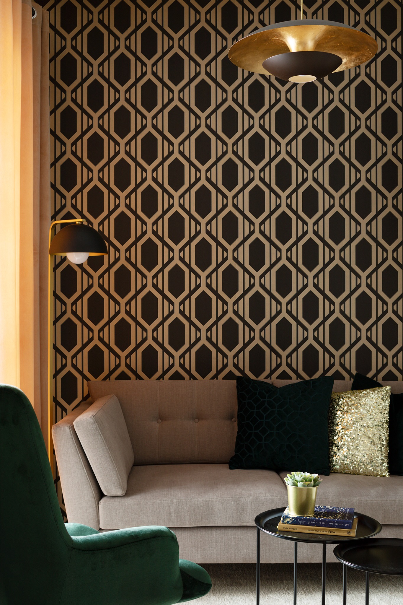 Fragment pokoju z geometryczną tapetą w odcieniach beżu i brązu, beżowa sofa z zieloną i złotą poduszką, zielony fotel, stolik kawowy z rośliną i książkami, złota lampa podłogowa i sufitowa.