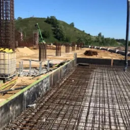 Siatka zbrojeniowa fundamentu na placu budowy, widoczne pionowe pręty zbrojeniowe i elementy szalunku, w tle zielone wzgórze i błękitne niebo.