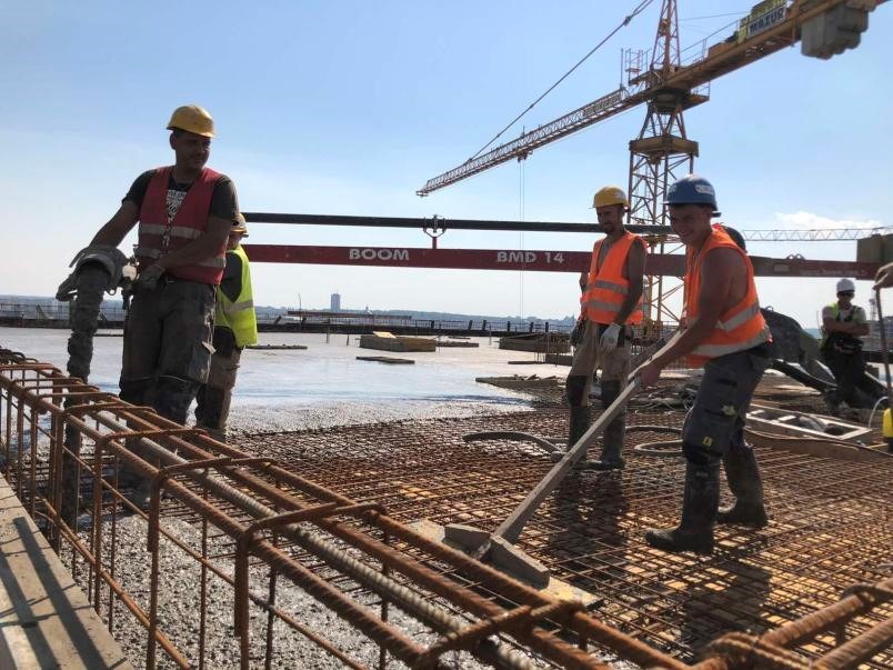 Robotnicy w kaskach i kamizelkach wylewają beton na zbrojenie na budowie budynku żelbetowego, z widocznym dźwigiem w tle.