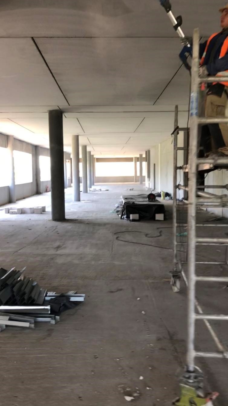 Wnętrze budynku w trakcie wykańczania sufitu podwieszanego, widoczny pracownik na drabinie montujący elementy konstrukcyjne, kolumny nośne oraz materiały budowlane na podłodze.