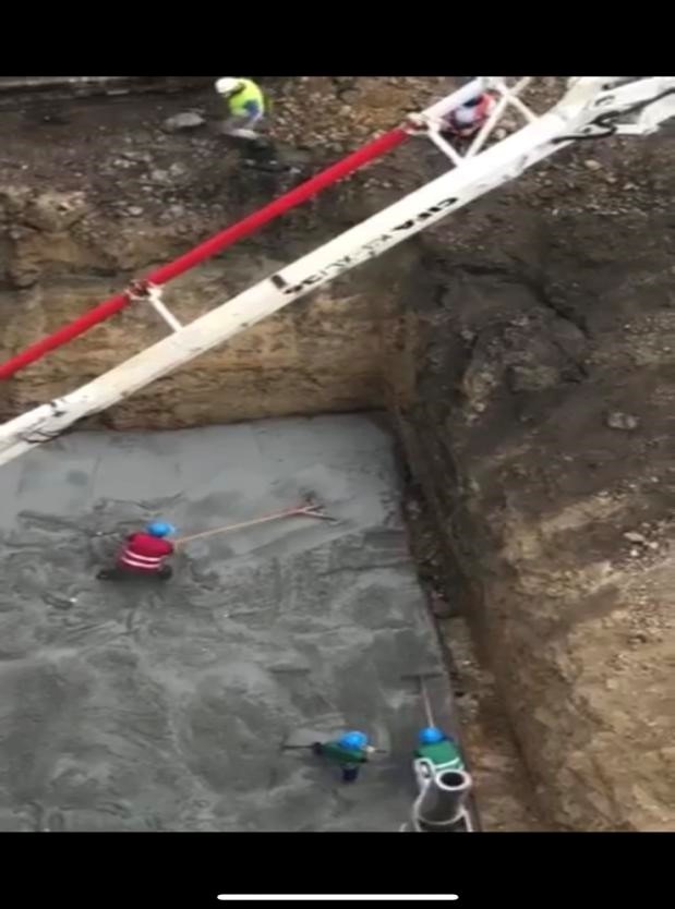Wylany fundament z betonu wyrównywany przez trzech pracowników w kaskach, z użyciem długich narzędzi, z pompy do betonu z czerwoną rurą, widok z góry.