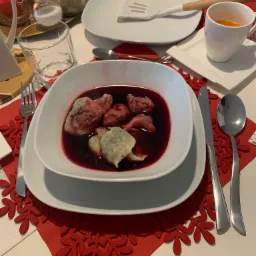 Uroczysty stół nakryty do obiadu, z talerzem czerwonego barszczu z uszkami na pierwszym planie, czerwone podkładki w kształcie płatków śniegu, w tle sałatka i przyprawy.