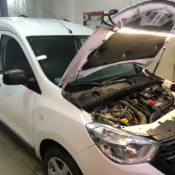 Biały samochód dostawczy z otwartą maską w warsztacie, widoczny silnik oświetlony lampą warsztatową, na ścianie schematy i zegar.