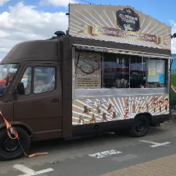 Brunatny food truck z reklamą 'Piekarnia Lodziarnia Cukiernia Węgierska', oferujący langosze, ciastka i gofry bąbelkowe, zaparkowany na brukowanym placu.