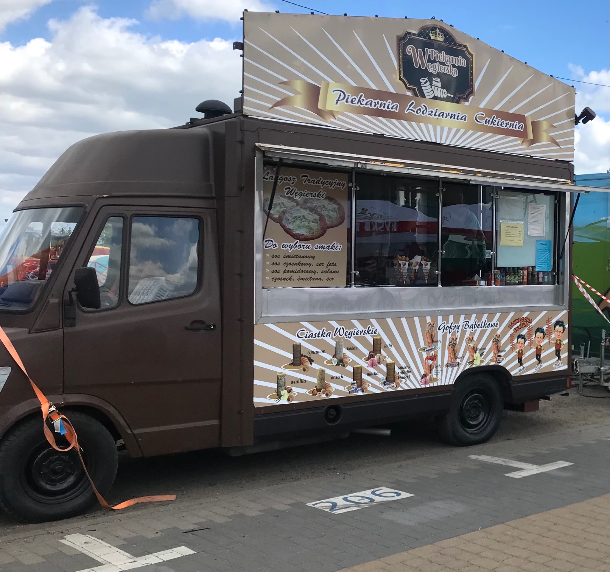 Brunatny food truck z reklamą 'Piekarnia Lodziarnia Cukiernia Węgierska', oferujący langosze, ciastka i gofry bąbelkowe, zaparkowany na brukowanym placu.