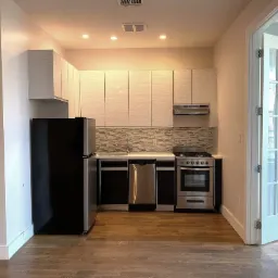 Wnętrze kuchni z białymi szafkami z poziomym wzorem, czarnymi dolnymi szafkami, stalowym zlewem, szarą mozaiką na ścianie, czarną lodówką i drewnianą podłogą, widok na otwarte, przeszklone drzwi.