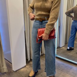 Zarini.jul - Osoba w przymierzalni prezentuje stylizację: beżowy sweter z włosem, szerokie jeansy z brązowym paskiem, beżowe buty na grubej podeszwie i czerwona torebka na ramieniu.