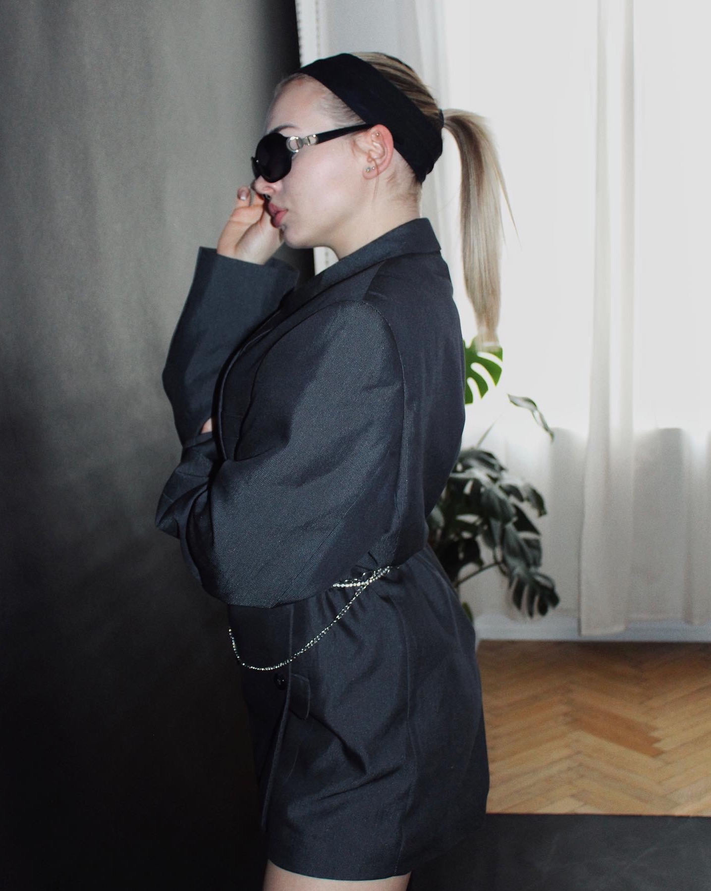 Kobieta z blond włosami spiętymi w kucyk, w czarnej opasce, okularach przeciwsłonecznych i ciemnej marynarce przewiązanej łańcuszkiem, pozuje na tle szarej ściany i okna z białą zasłoną.