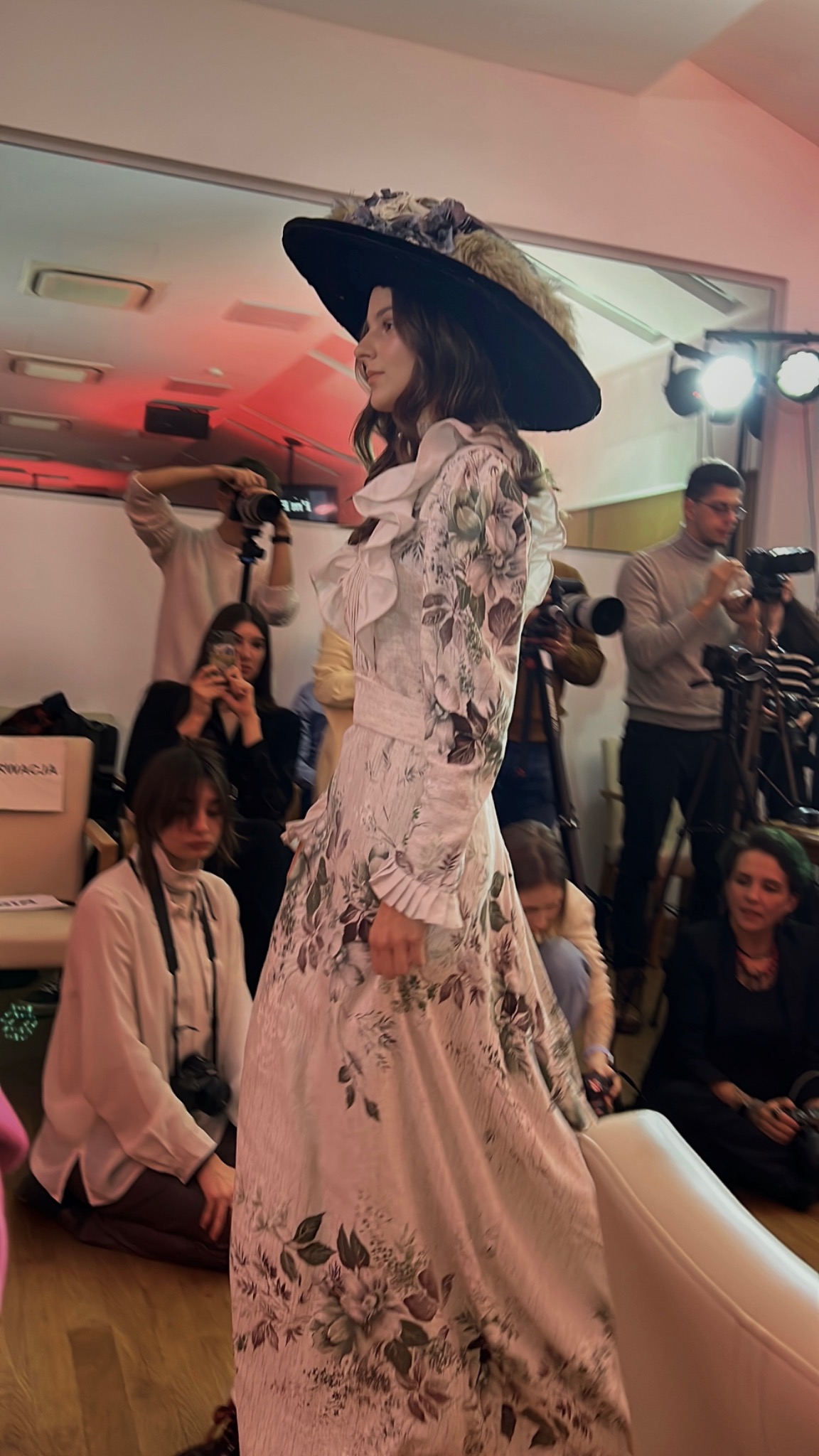 Modelka prezentuje suknię w stylu retro z kwiatowym wzorem i dużym kapeluszem na pokazie mody, otoczona fotografami.