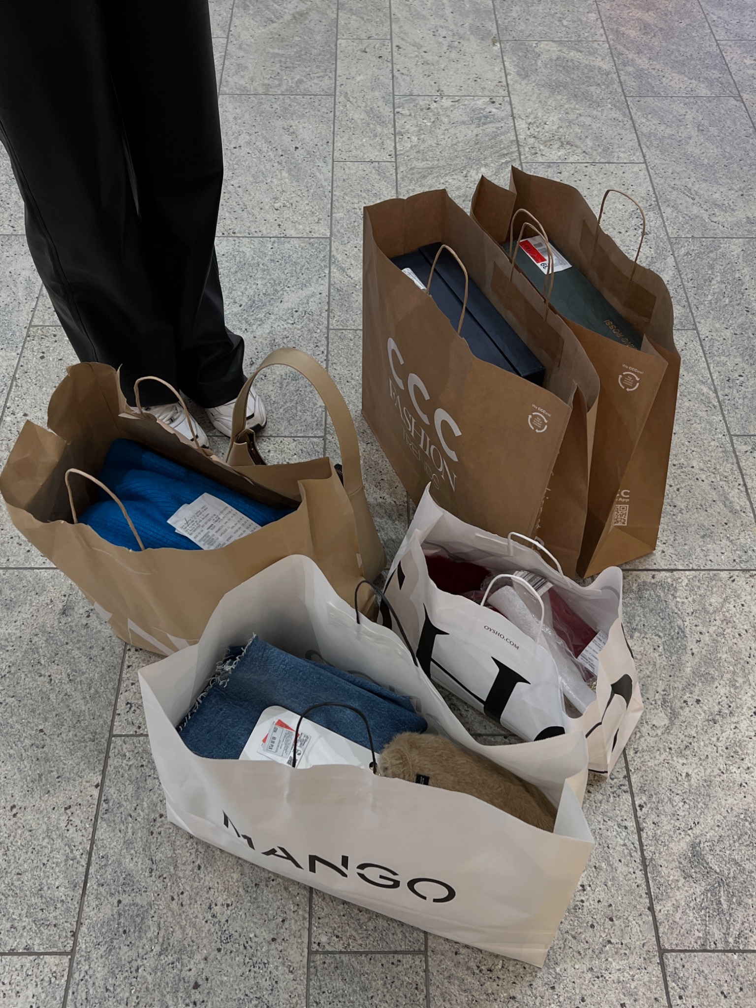 Zakupy w torbach z logo CCC, Oysho i Mango leżące na podłodze z płytek, obok osoby w czarnych spodniach i białych butach sportowych.