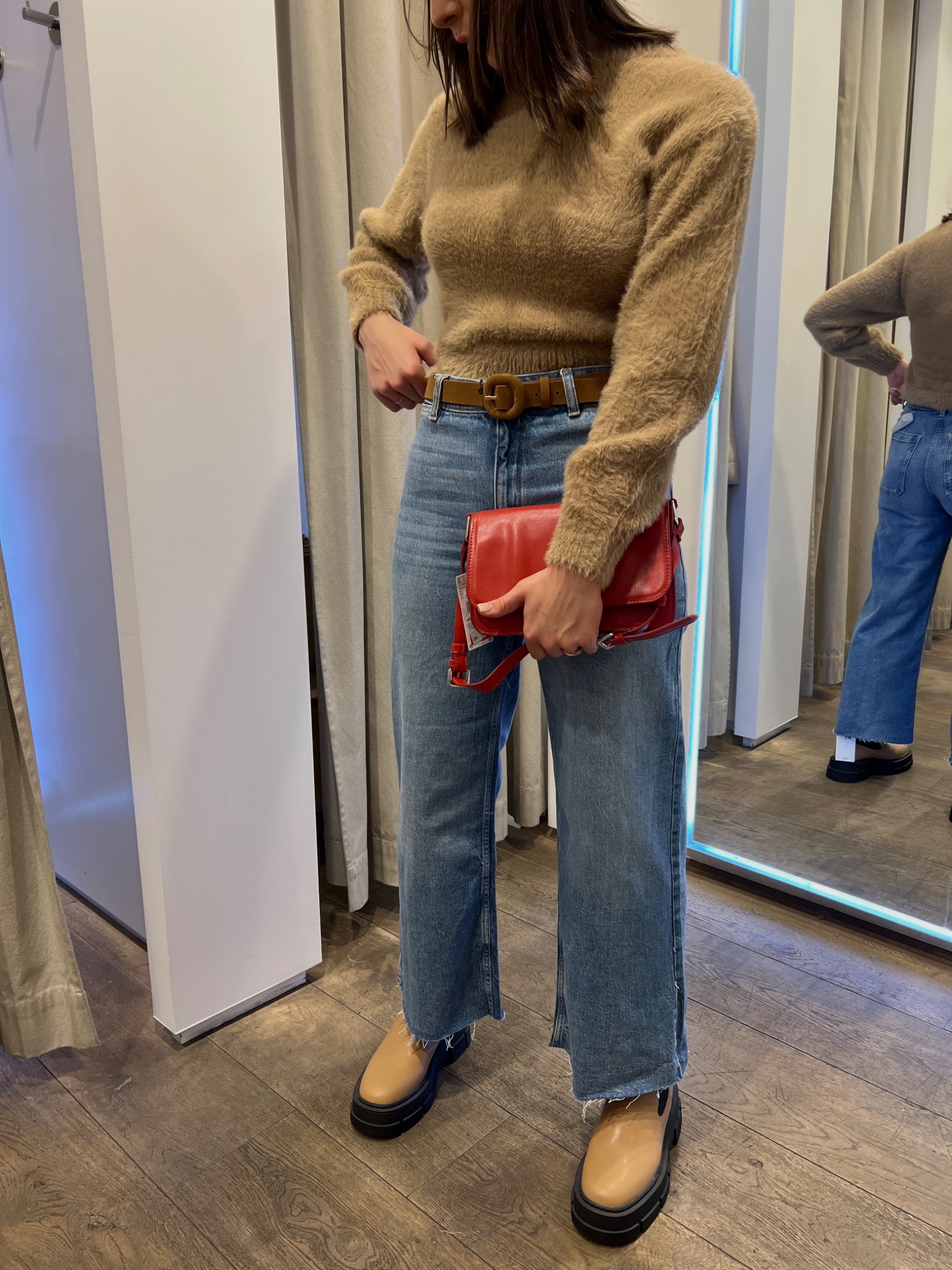 Osoba w przymierzalni prezentuje stylizację: beżowy sweter z włosem, szerokie jeansy z brązowym paskiem, beżowe buty na grubej podeszwie i czerwona torebka na ramieniu.