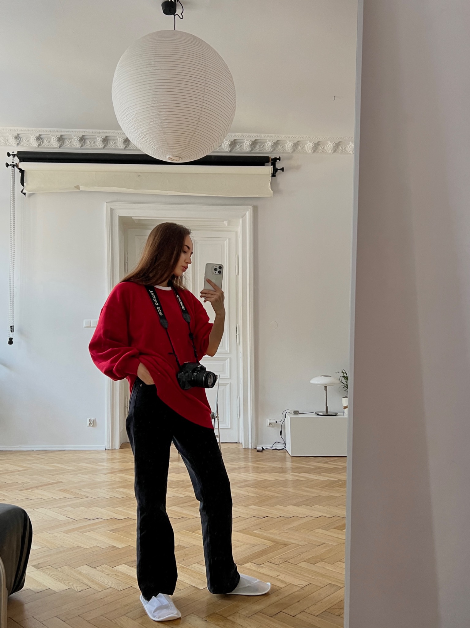 Kobieta robi selfie w lustrze, ubrana w czerwoną bluzę i czarne spodnie, z aparatem fotograficznym na szyi, widoczny parkiet i minimalistyczny wystrój wnętrza.