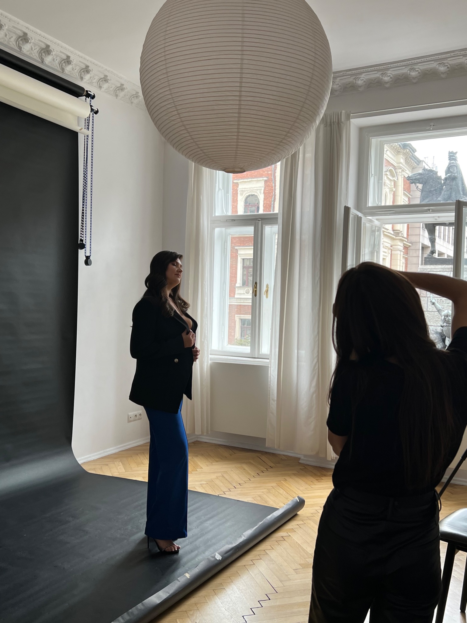 Sesja zdjęciowa w studio: kobieta w czarnej marynarce i niebieskich spodniach pozuje na tle czarnego tła, fotograf z tyłu kadru, widok na rzeźbę za oknem.