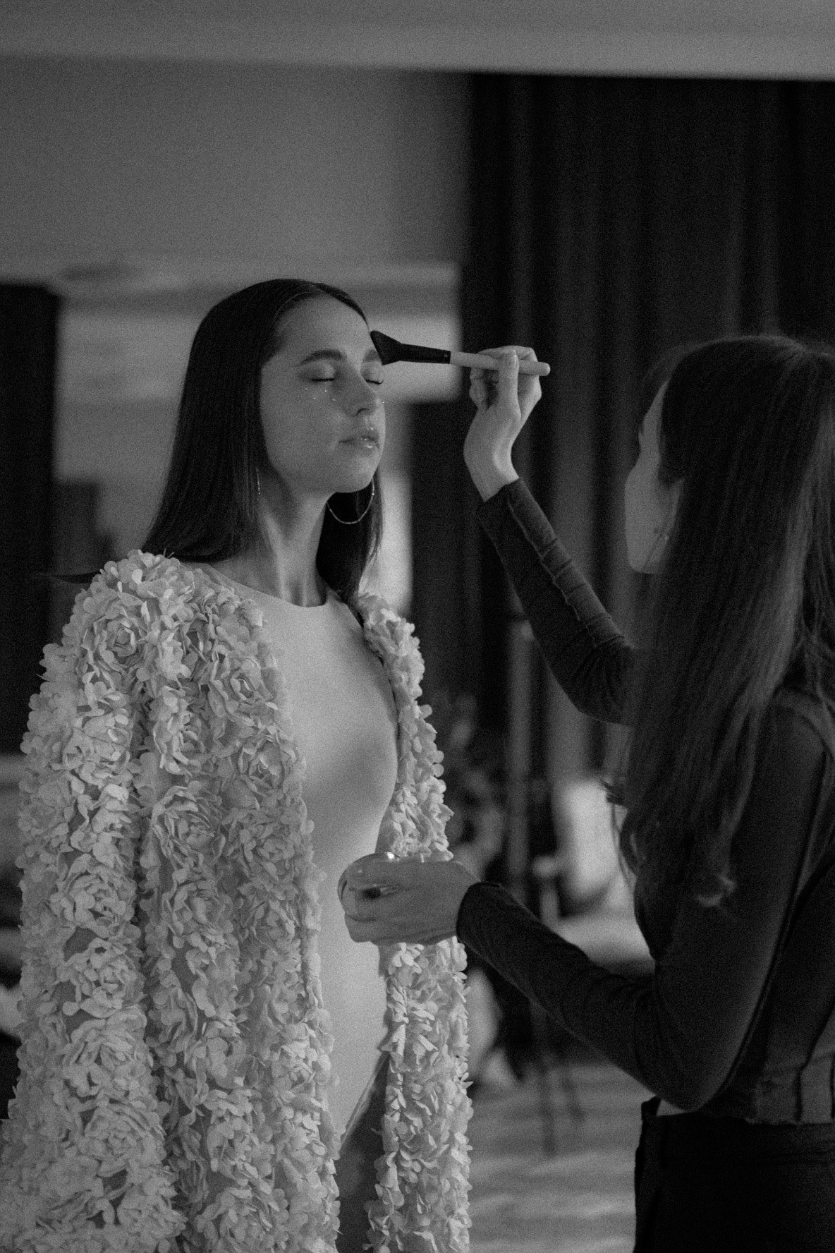 Czarno-białe zdjęcie: Stylistka aplikuje pędzlem makijaż na twarz modelki z zamkniętymi oczami, noszącej ozdobny żakiet w kwiaty i duże kolczyki koła.