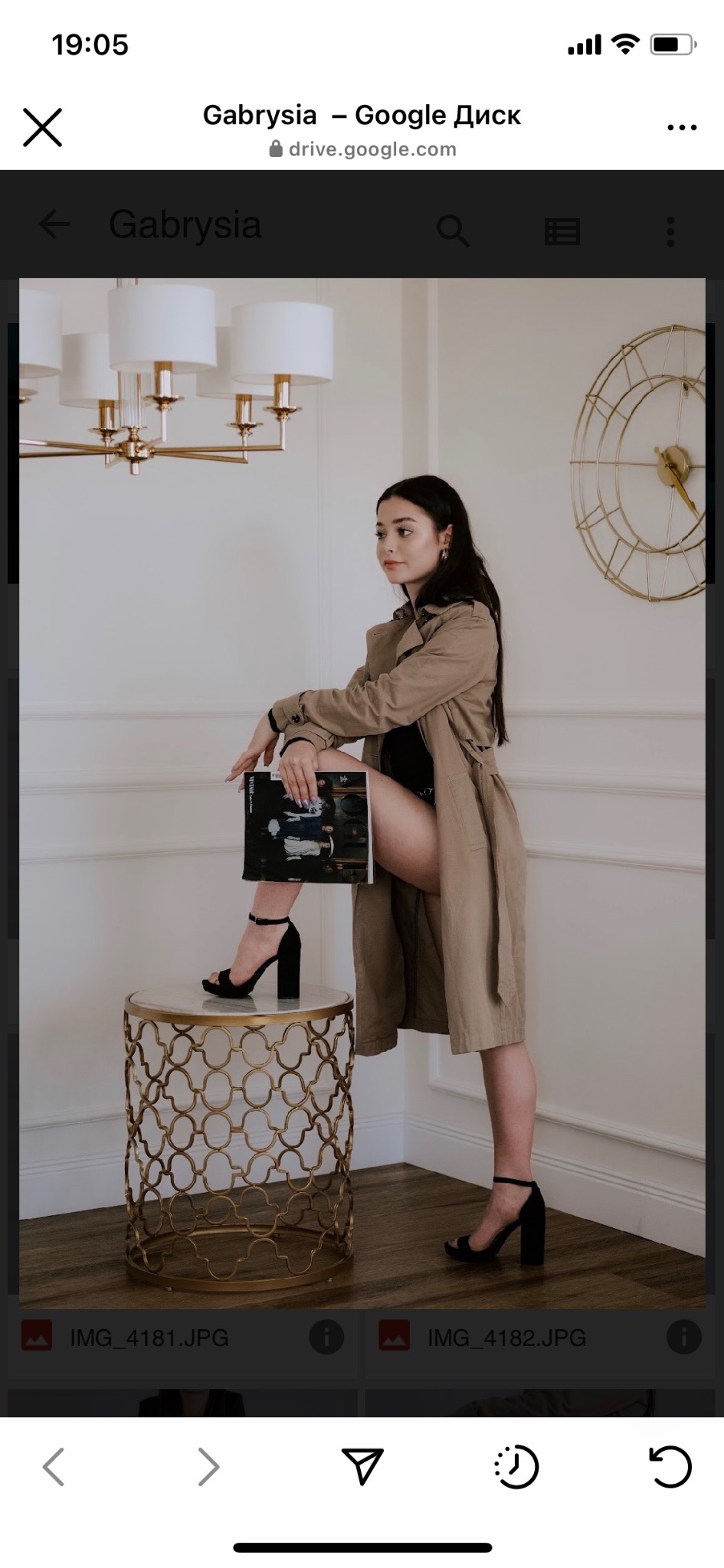 Kobieta w płaszczu pozuje z nogą opartą na złotym stoliku, trzymając magazyn modowy w eleganckim wnętrzu.