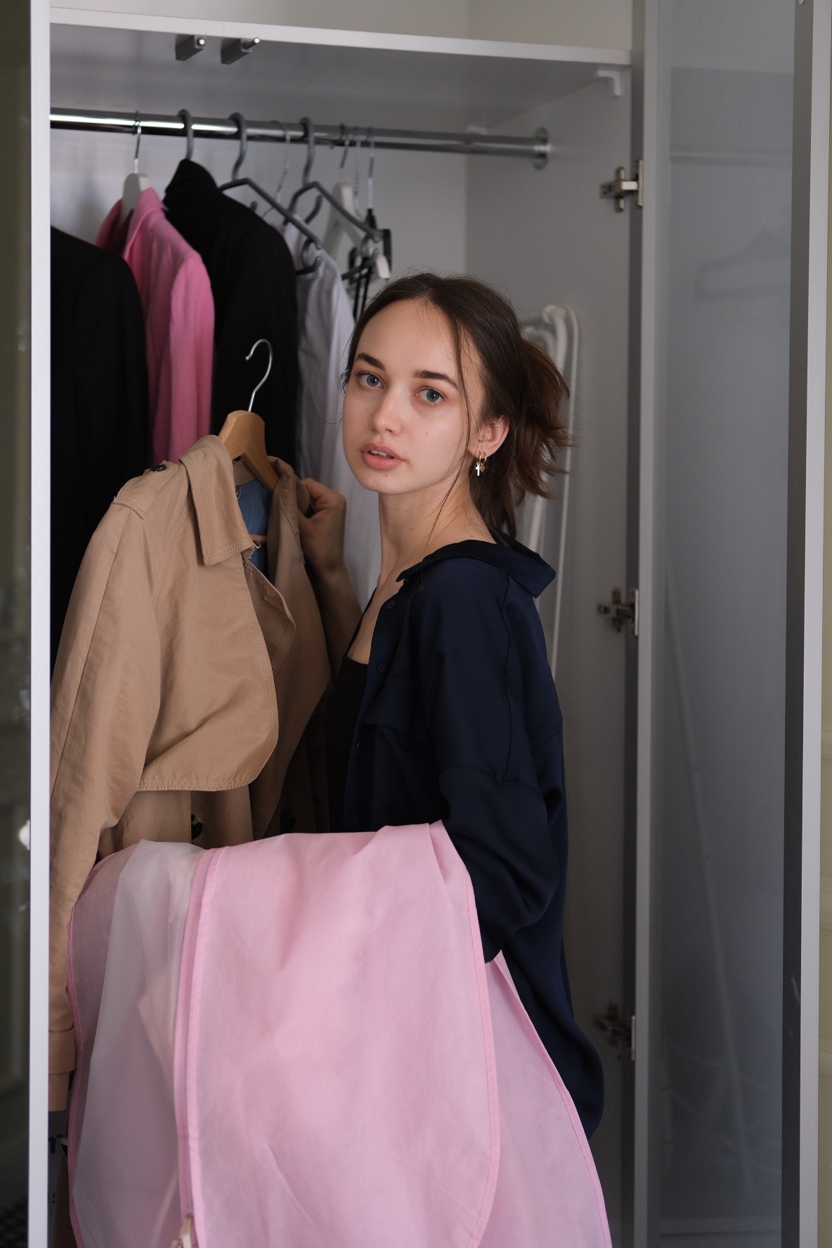 Młoda kobieta w granatowej bluzie, stojąca przed otwartą szafą z ubraniami, trzyma beżowy płaszcz na wieszaku i różowy pokrowiec na ubrania.