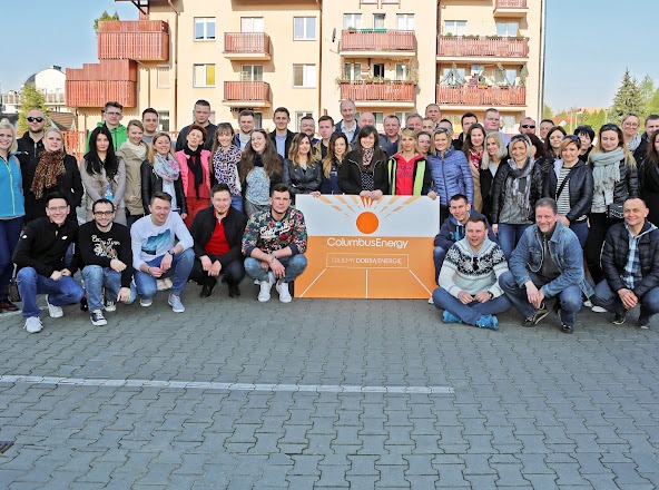 Zespół Columbus Energy pozuje z pomarańczowym banerem przed budynkiem mieszkalnym.