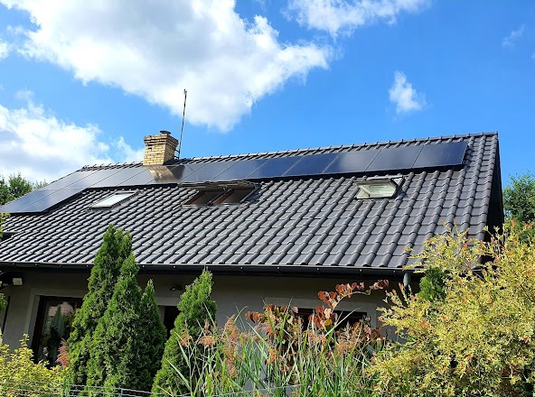 Czarny panel słoneczny na ciemnoszarym dachu domu jednorodzinnego z ceglanym kominem i oknami dachowymi, widoczne iglaki i krzewy w ogrodzie, błękitne niebo z chmurami w tle.