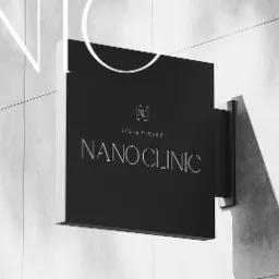 Czarny szyld NanoClinic z minimalistycznym logo i napisem Julia Piejuć na jasnej, kafelkowej ścianie, uchwycony pod kątem.