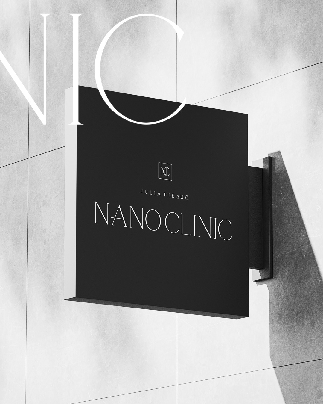Czarny szyld NanoClinic z minimalistycznym logo i napisem Julia Piejuć na jasnej, kafelkowej ścianie, uchwycony pod kątem.
