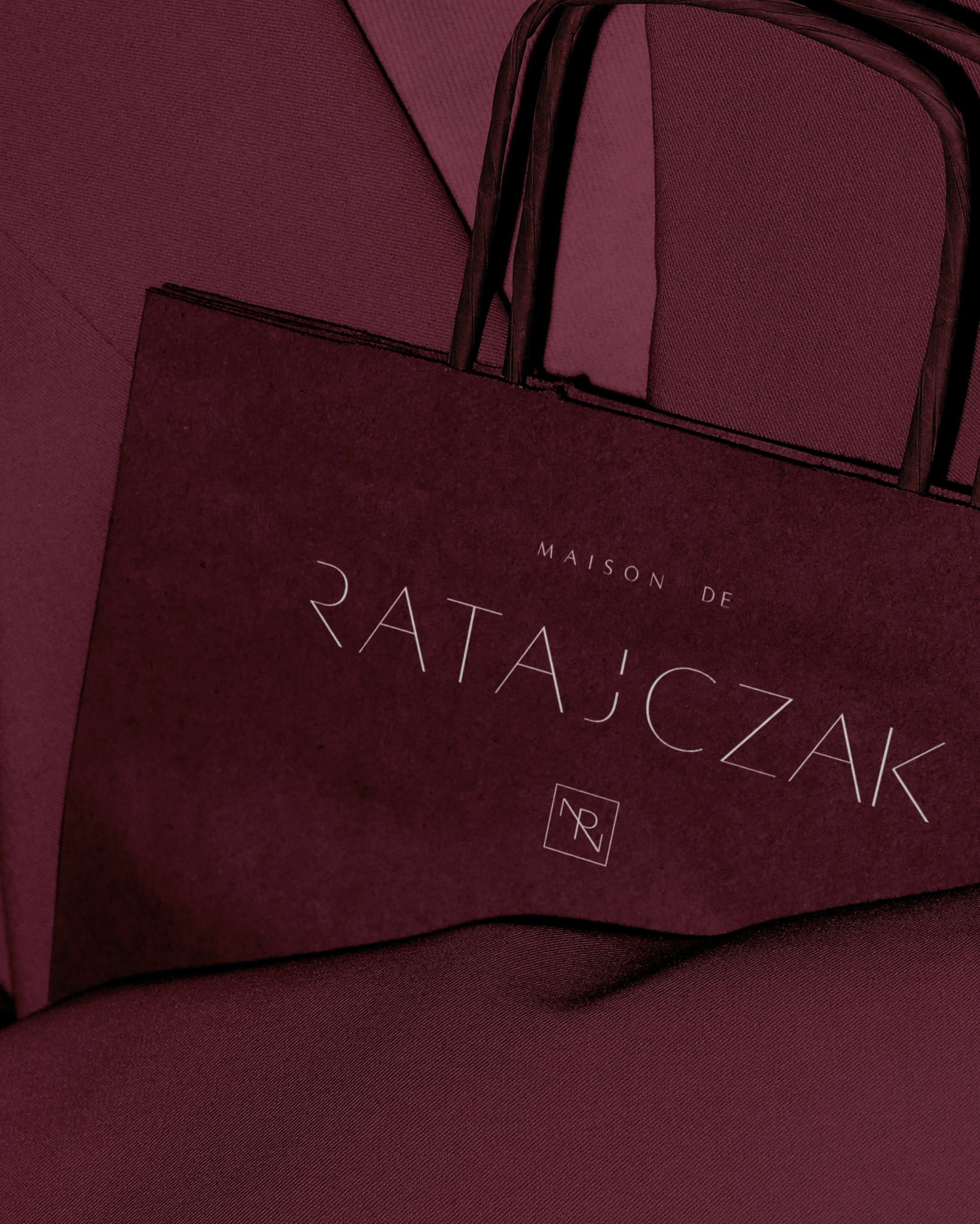 Bordowa torba papierowa z logo 'Maison de Ratajczak' na tle bordowej tkaniny. Minimalistyczny design, elegancki detal identyfikacji wizualnej marki.