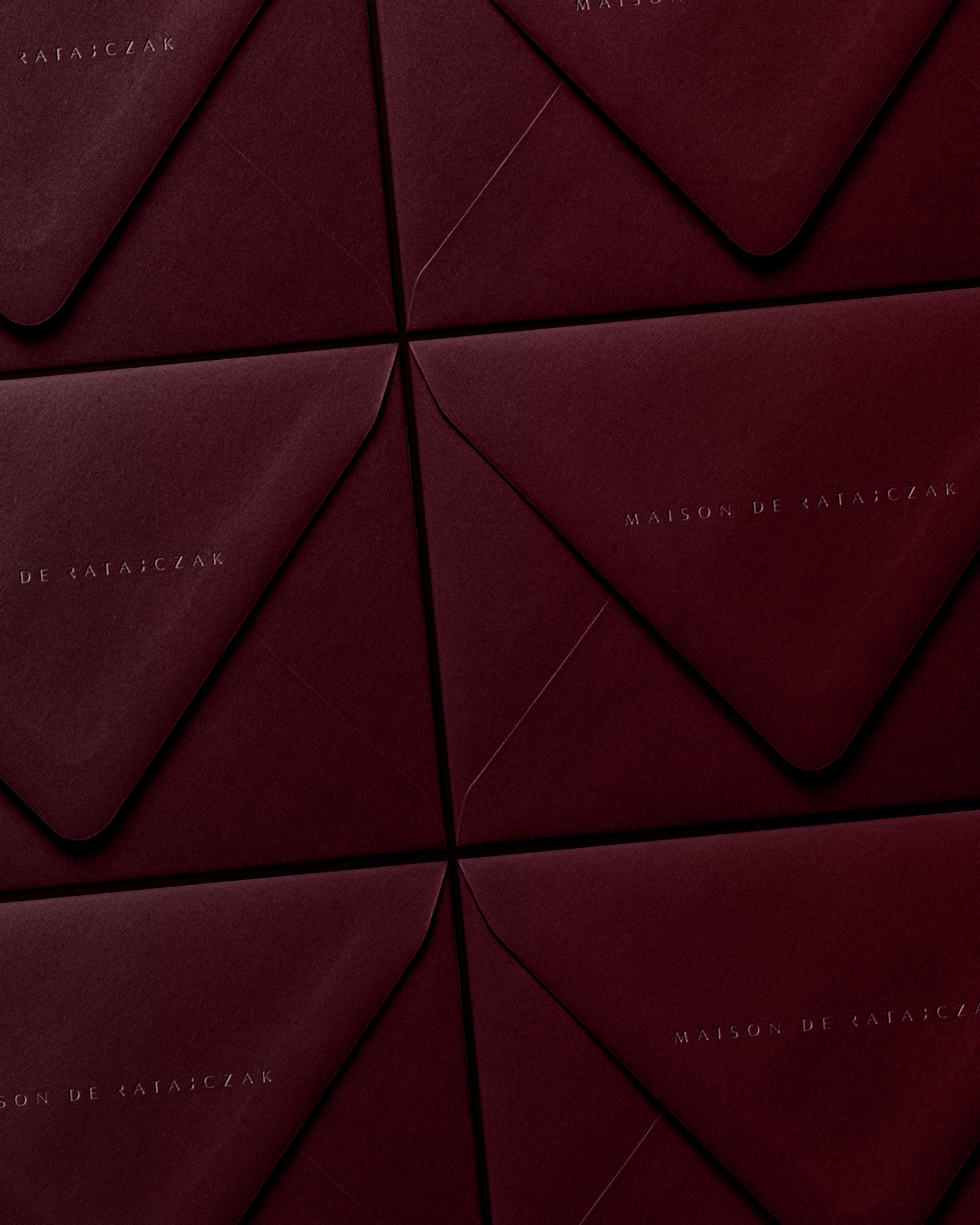 Eleganckie, bordowe koperty z subtelnym tłoczeniem logo 'Maison de Ratajczak' ułożone w geometryczny wzór. Minimalistyczny design, stonowana kolorystyka.