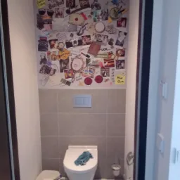 Kreatywna ściana w toalecie po remoncie w Pile, ozdobiona kolażem zdjęć i pamiątek, z widoczną szczotką do WC i pojemnikiem na narzędzia malarskie, sugerującym niedawne prace wykończeniowe.