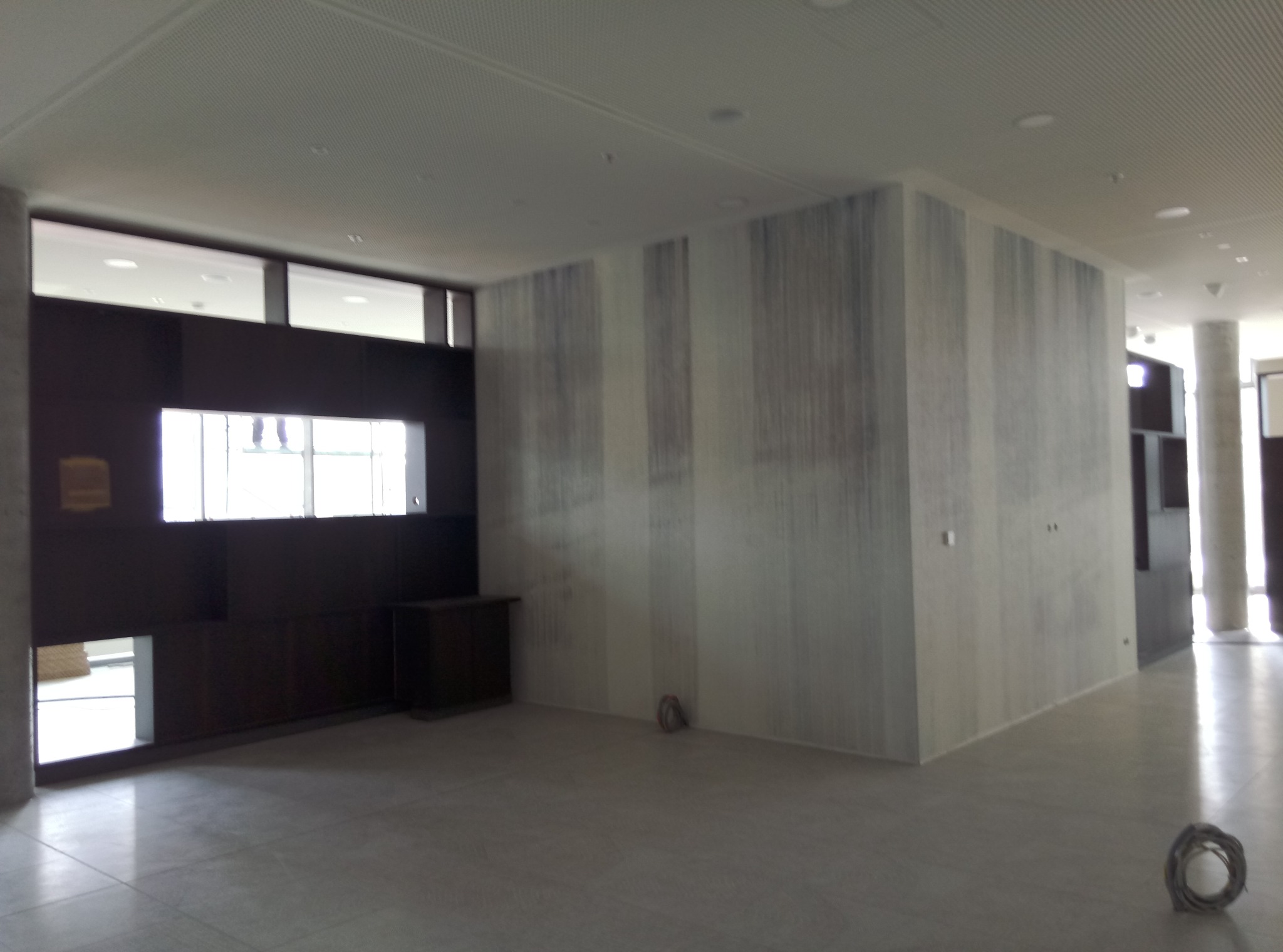 Wnętrze biurowe z nowymi ścianami w odcieniach szarości, przypominające efekt akwareli, widoczny kabel zwinięty w kłębek na podłodze, oraz fragment okna z sylwetkami osób w tle.