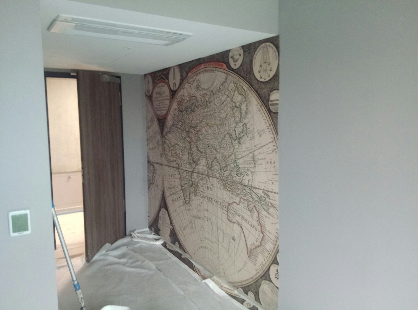 Tapeta z motywem mapy świata w stylu vintage, częściowo zakryta folią ochronną podczas montażu, widoczny fragment drzwi i sufitu.