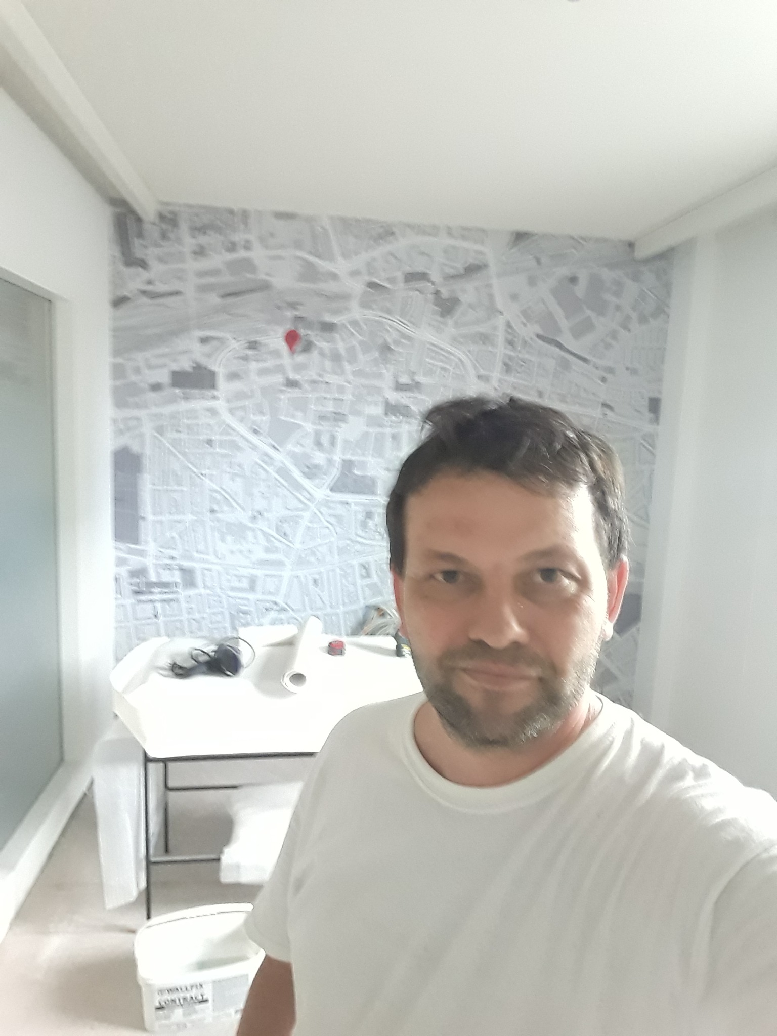 Mężczyzna w białej koszulce robi selfie na tle ściany z tapetą przedstawiającą plan miasta z czerwoną pinezką, widoczny stół z narzędziami i rolką tapety.
