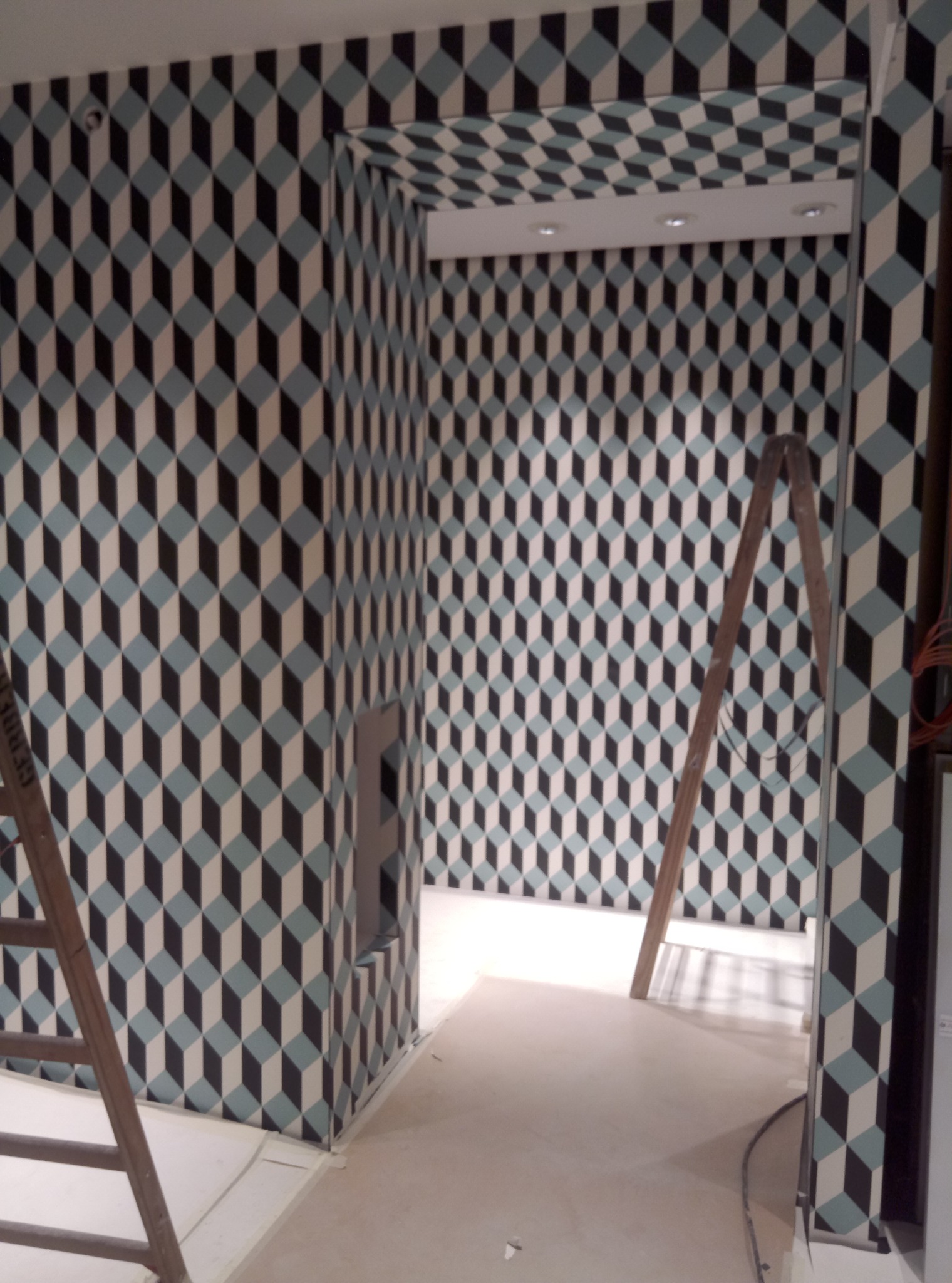 Wnętrze pokoju po remoncie, ściany i sufit pokryte tapetą w geometryczny wzór trójwymiarowych kostek w odcieniach bieli, błękitu i czerni, widoczna drewniana drabina oparta o ścianę, podłoga...