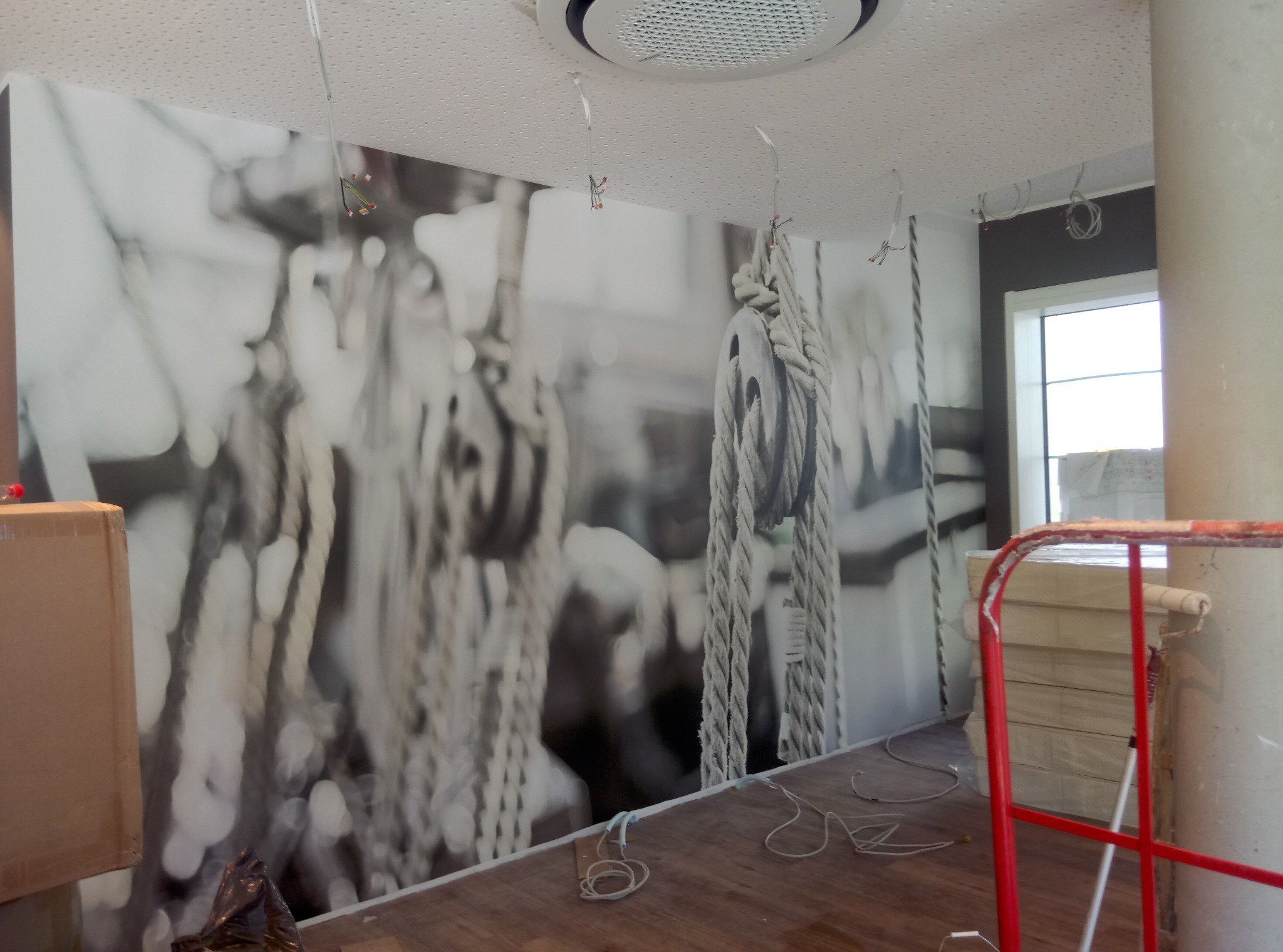 Duża, czarno-biała fototapeta z motywem żeglarskim (liny, bloczki) na ścianie w pomieszczeniu w trakcie remontu, widoczne narzędzia malarskie i materiały budowlane.