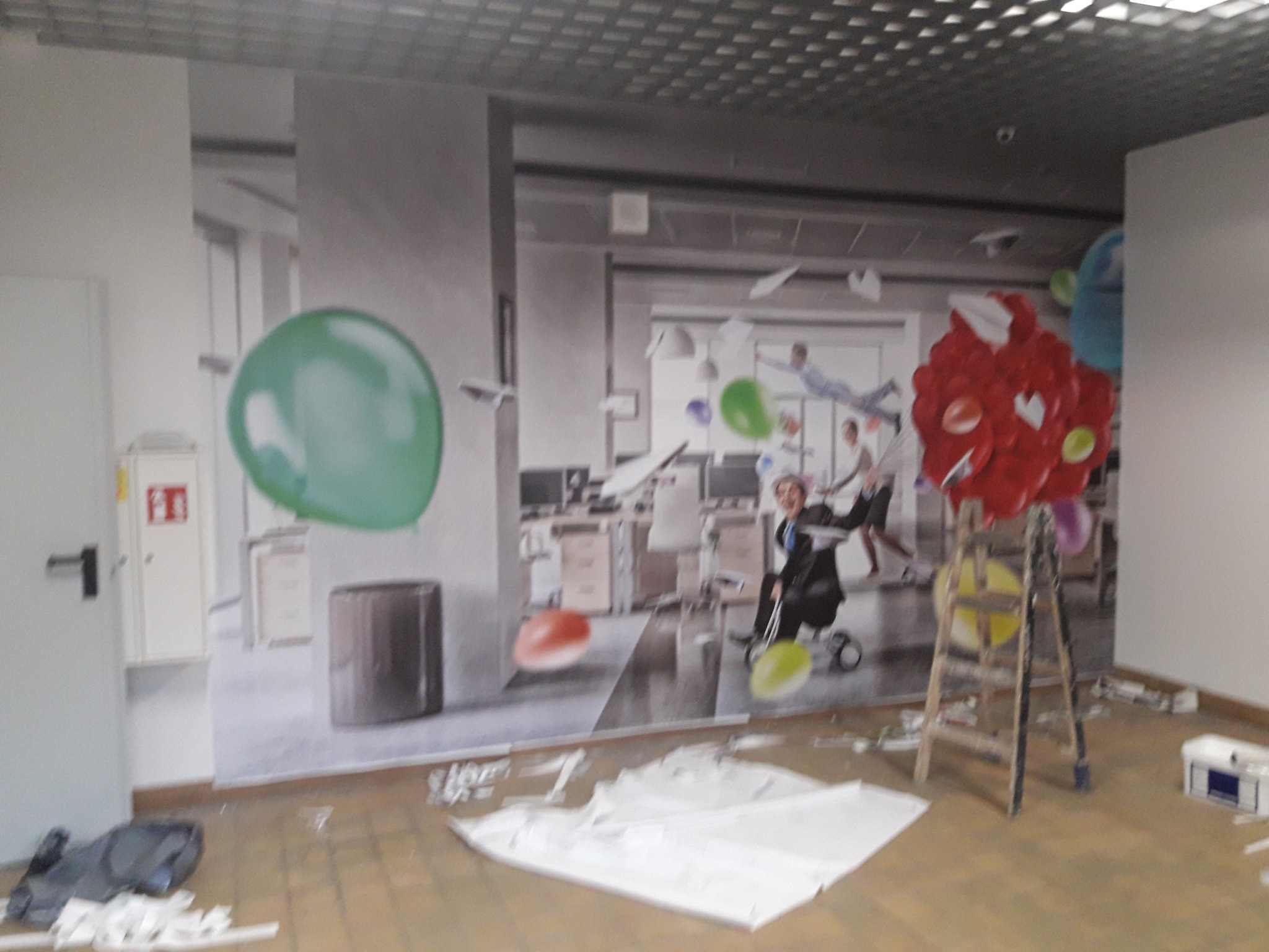 Świeżo położona fototapeta z motywem biura i balonów, widoczny fragment drabiny i resztki materiałów montażowych na podłodze.