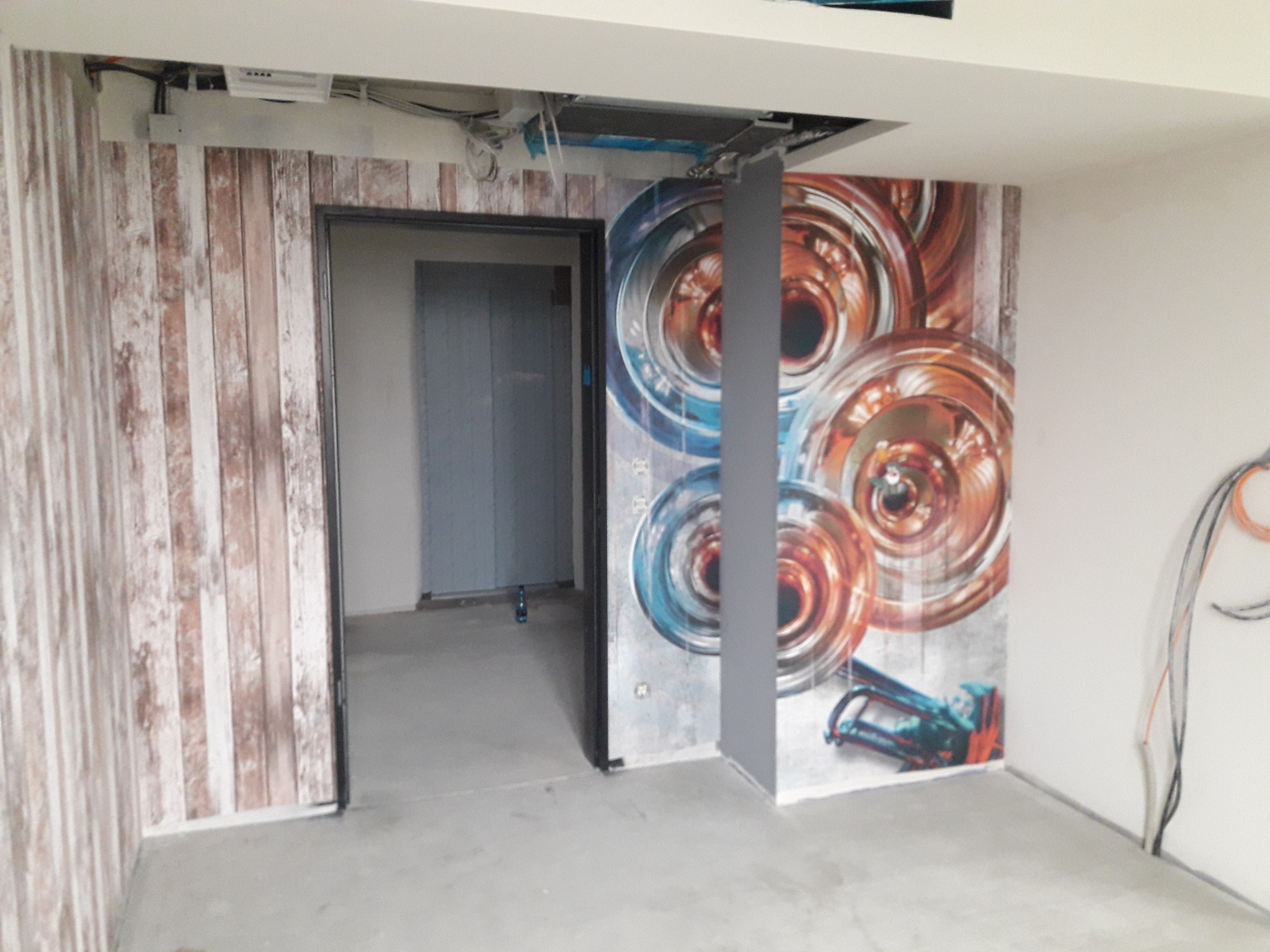 Wnętrze po remoncie: ściana z tapetą imitującą drewniane deski obok otwartych drzwi, druga ściana z abstrakcyjnym wzorem okręgów w odcieniach niebieskiego i pomarańczowego, widoczny zszywacz...