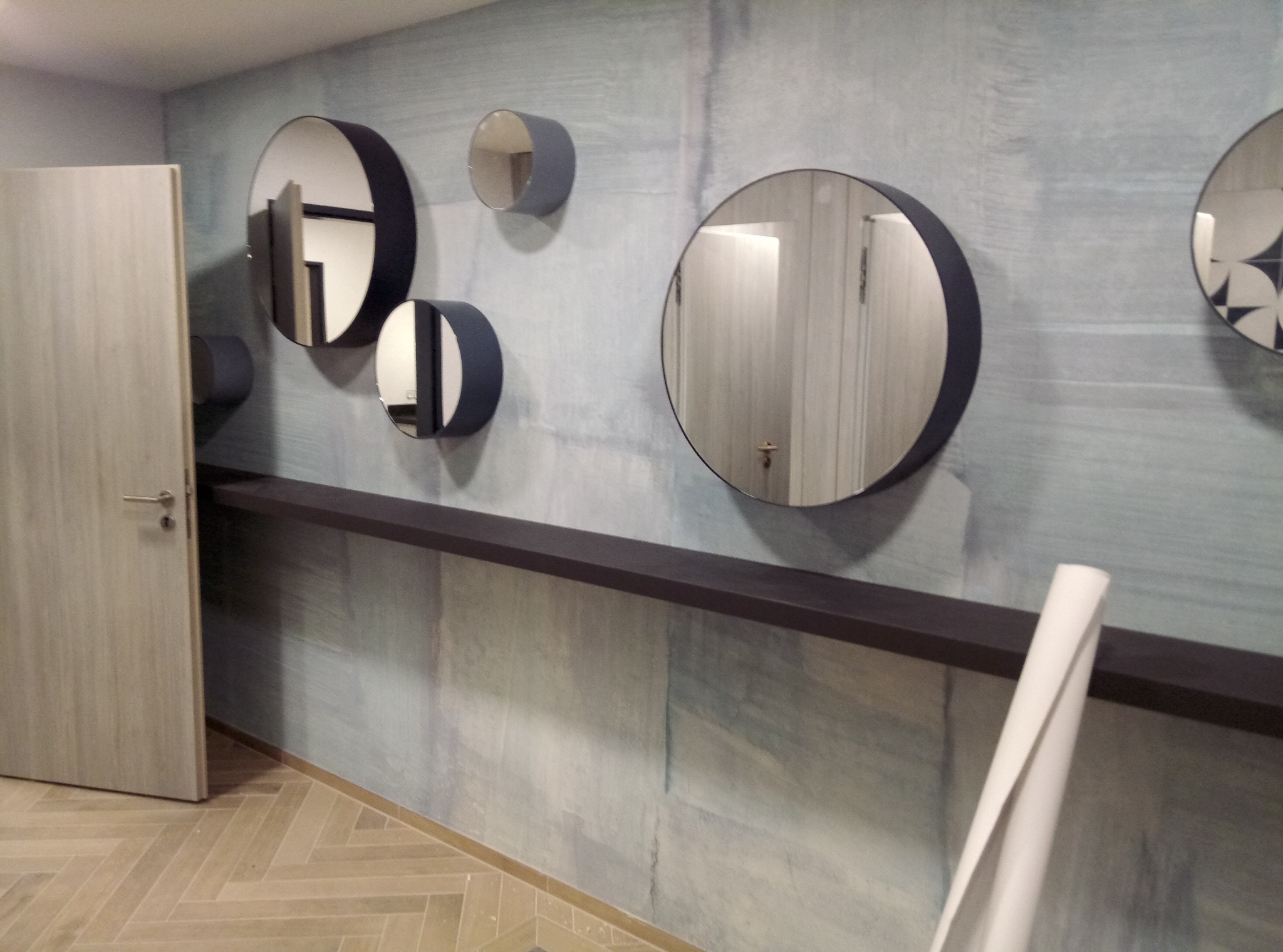 Ściana w odcieniach szarości z abstrakcyjnym wzorem przypominającym pociągnięcia pędzlem, ozdobiona okrągłymi lustrami w ciemnych ramkach i półką, na podłodze parkiet w jodełkę, widoczny fragment...