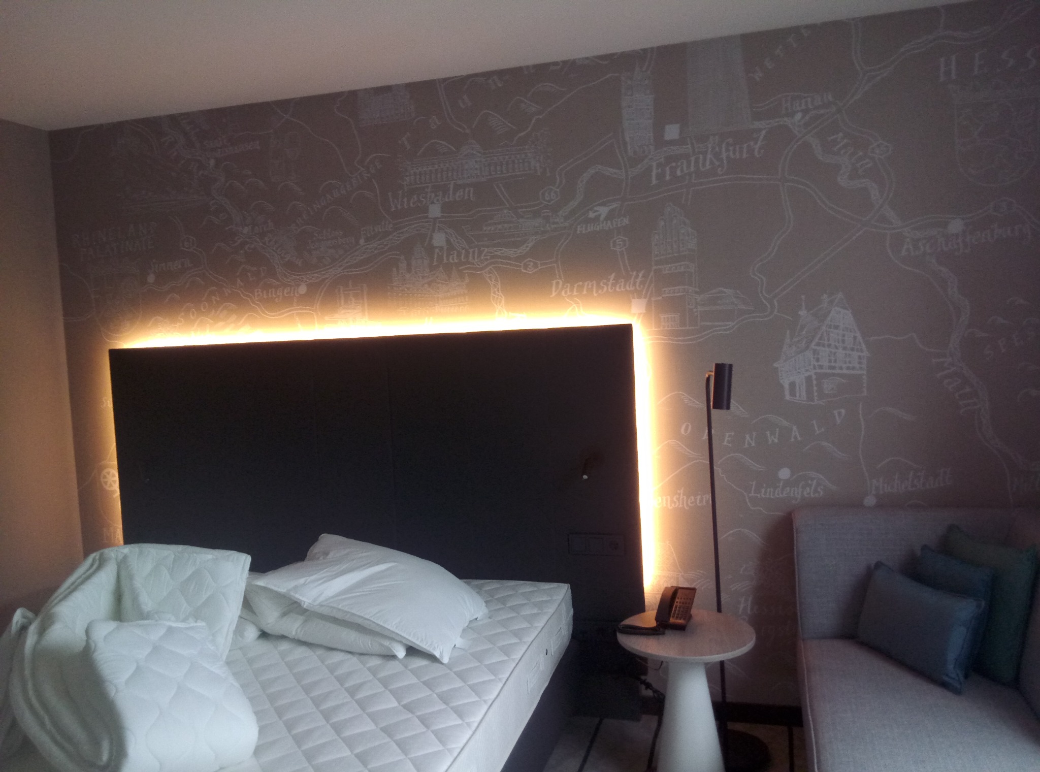 Nowoczesna sypialnia z podświetlanym zagłówkiem i tapetą w mapę miast Niemiec, w tym Frankfurtu i Wiesbaden.