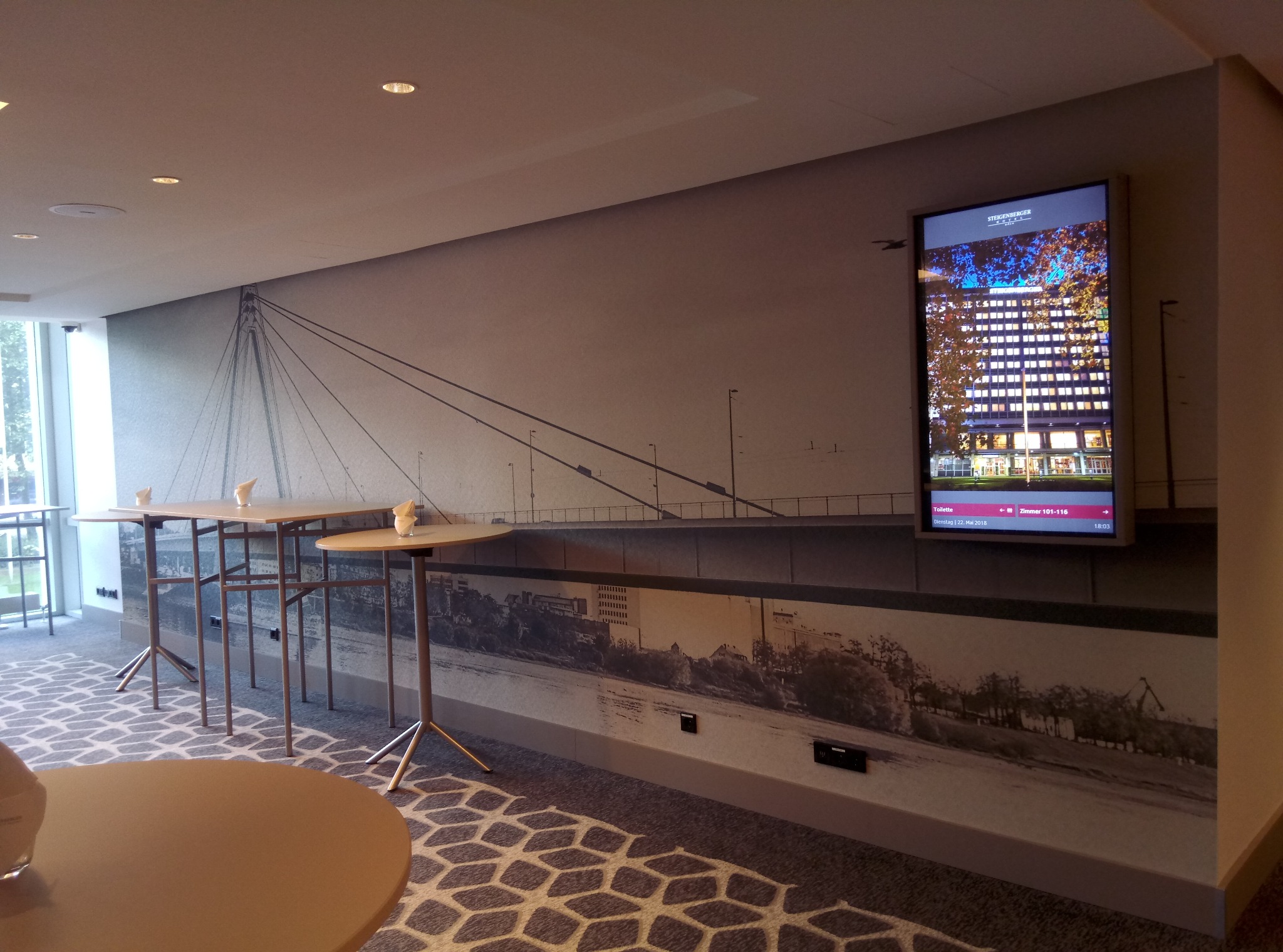 Wnętrze sali konferencyjnej z fototapetą przedstawiającą most i miasto, uzupełnioną o ekran reklamowy Steigenberger Hotel. Wysokie stoliki koktajlowe z jasnego drewna na trójnożnych metalowych nogach.