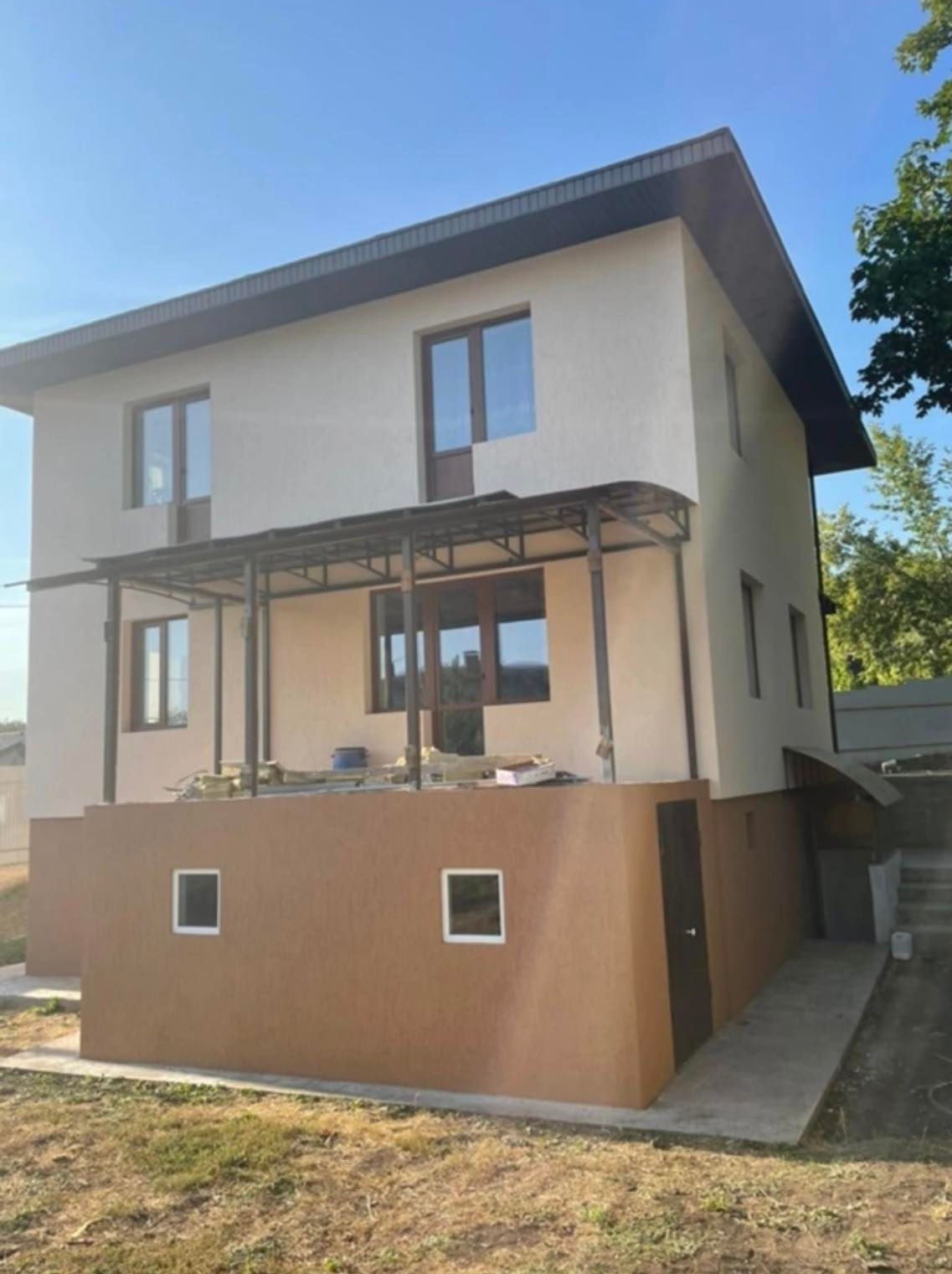 Dwukondygnacyjny dom z jasną elewacją i brązowym cokołem, widoczna zadaszona weranda z metalową konstrukcją, trawnik przed budynkiem.