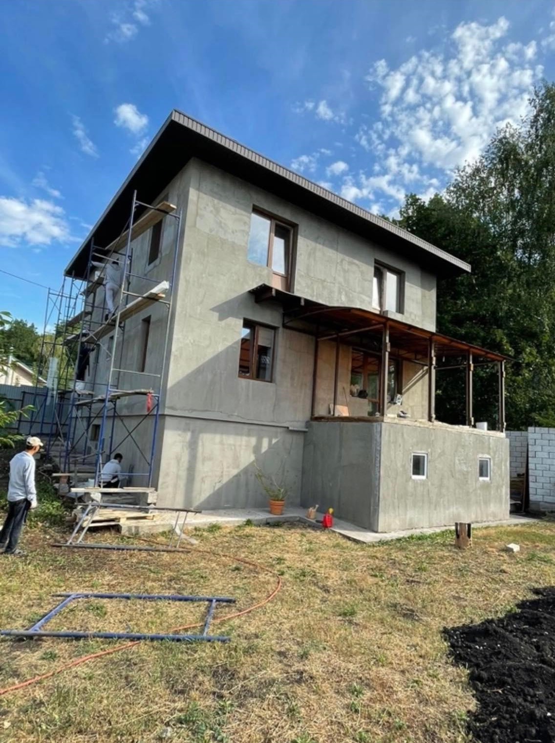 Dwukondygnacyjny dom w trakcie remontu elewacji, widoczne rusztowania i pracownicy, surowy tynk, niezadaszony taras i fragment ogrodu.