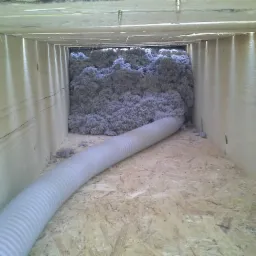 Wnętrze drewnianej konstrukcji z widoczną warstwą szarej izolacji w postaci włókniny i wężem do jej rozprowadzania, leżącym na podłodze z płyt OSB.