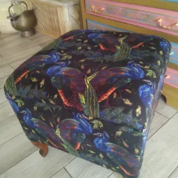 Kwadratowa pufa z podnoszonym siedziskiem, obita czarną tkaniną w motywy pawich piór, stoi na drewnianej podłodze w jasnym odcieniu, w tle widoczna komoda w pastelowych kolorach z pozłacanymi...