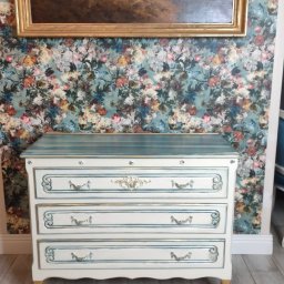 MirroProject - Biała komoda z trzema szufladami, zdobiona niebieskimi i złotymi detalami, stoi na tle ściany w kwiatowy wzór i obrazu w złotej ramie.