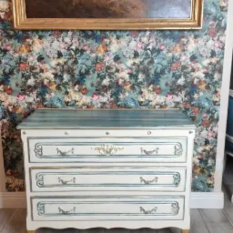 Biała komoda z trzema szufladami, zdobiona niebieskimi i złotymi detalami, stoi na tle ściany w kwiatowy wzór i obrazu w złotej ramie.