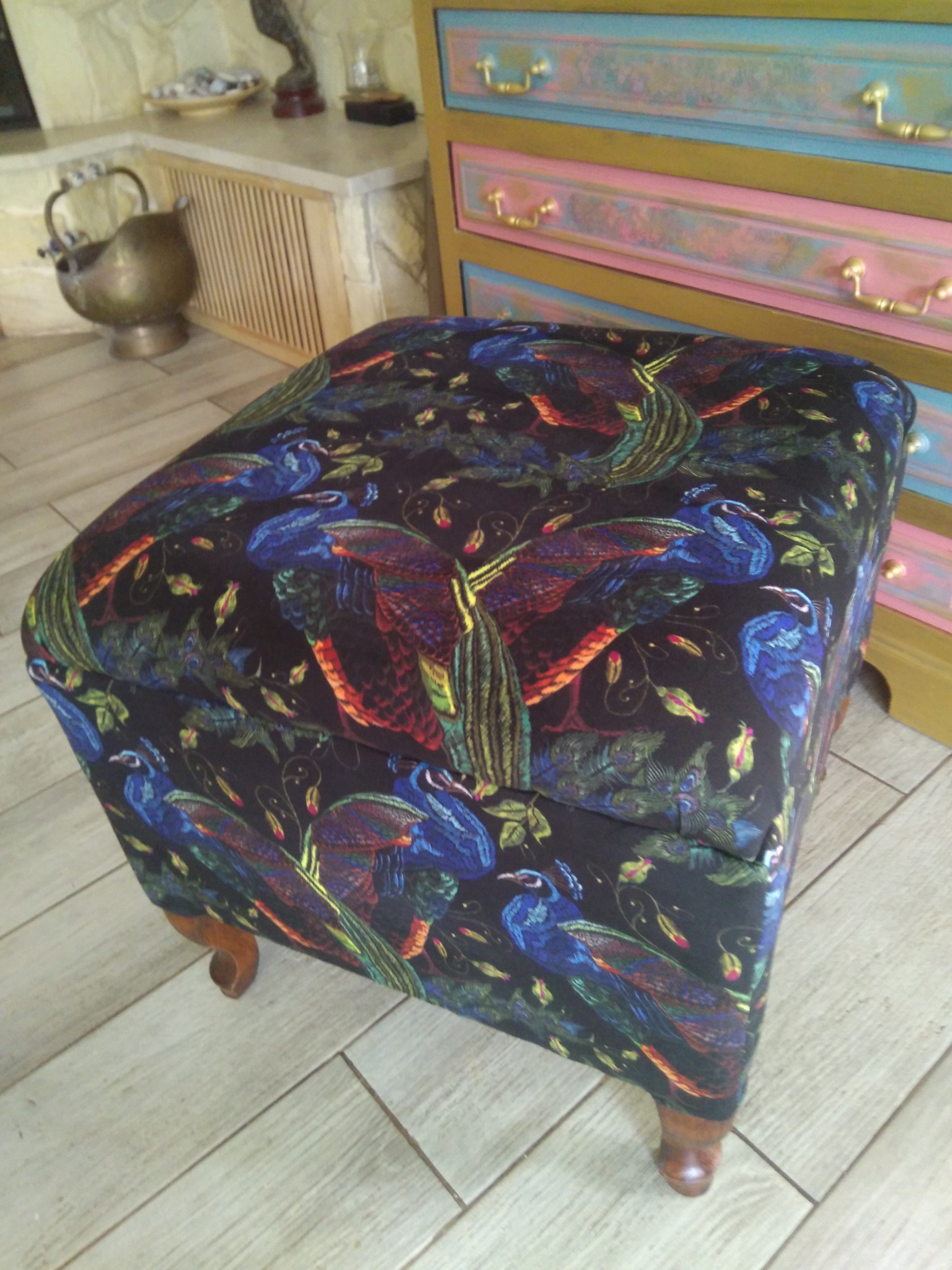 Kwadratowa pufa z podnoszonym siedziskiem, obita czarną tkaniną w motywy pawich piór, stoi na drewnianej podłodze w jasnym odcieniu, w tle widoczna komoda w pastelowych kolorach z pozłacanymi...