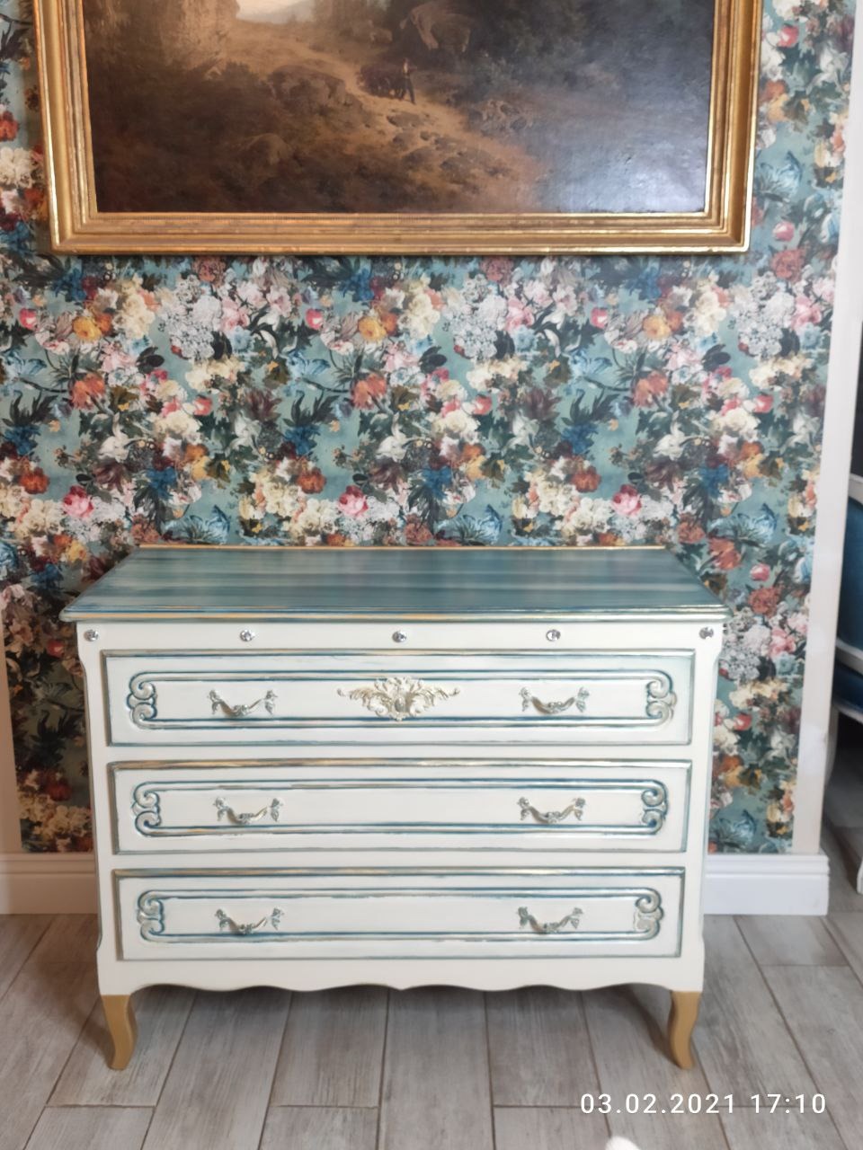 Biała komoda z trzema szufladami, zdobiona niebieskimi i złotymi detalami, stoi na tle ściany w kwiatowy wzór i obrazu w złotej ramie.