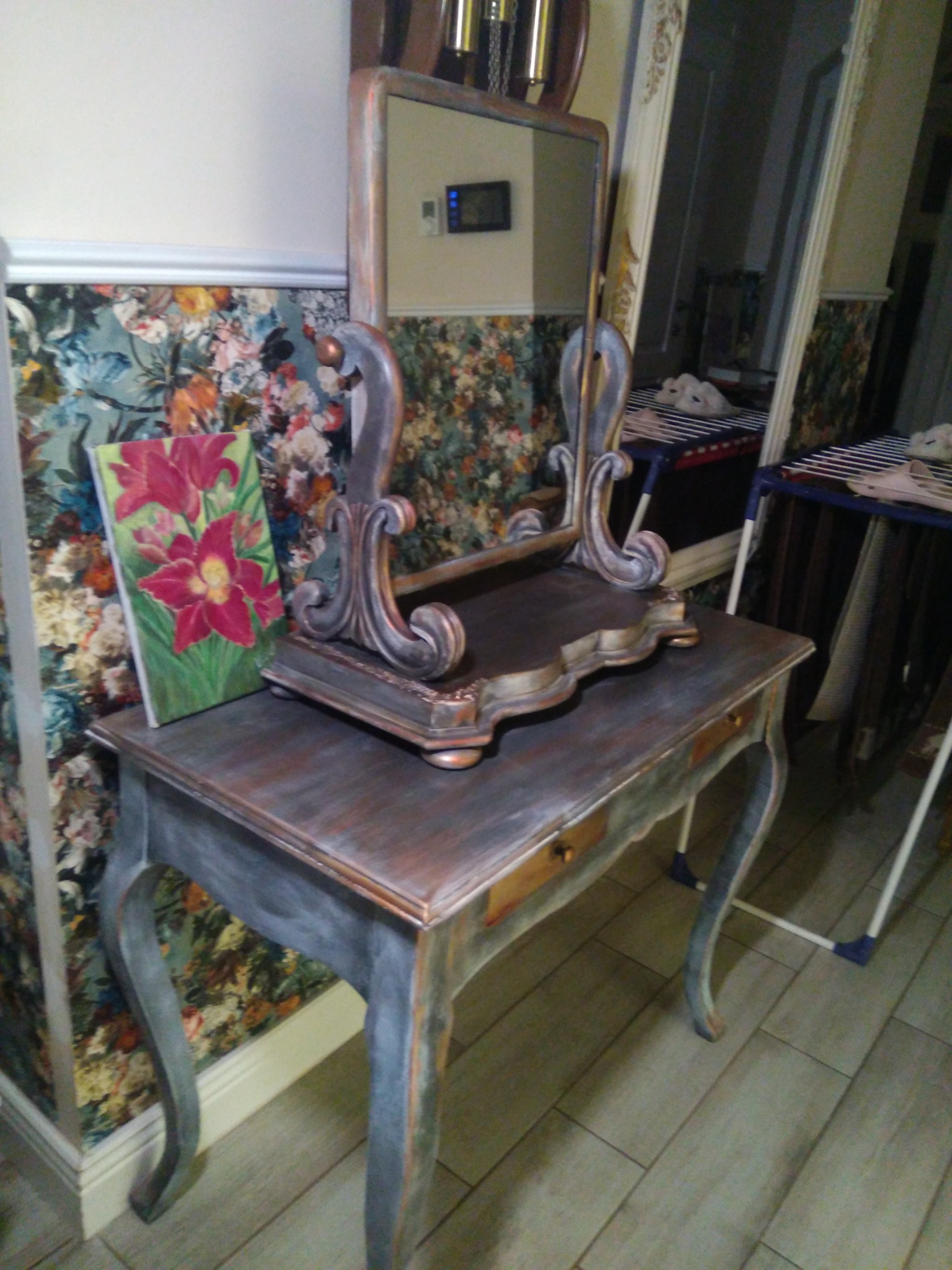 Stylizowana toaletka w odcieniach szarości i brązu z lustrem i obrazem z motywem kwiatowym na blacie, ustawiona na tle ściany z tapetą w kwiaty, w pomieszczeniu z wyłożoną podłogą.